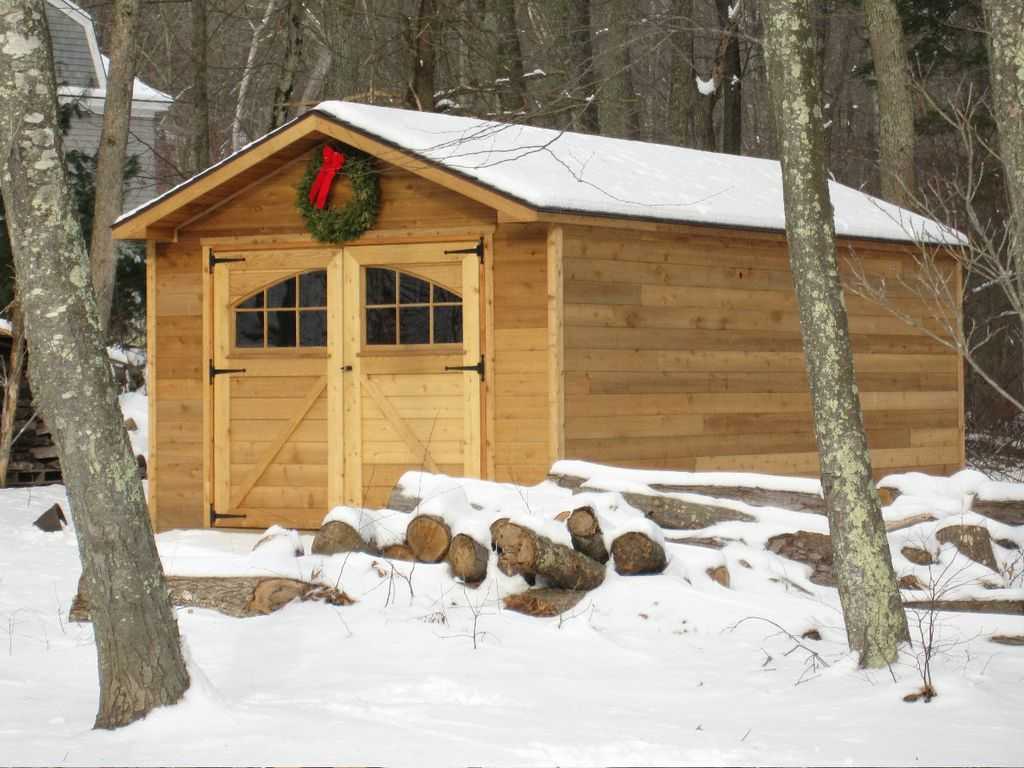 Peak-style wood-sided storage shed with double doors and transom windows, decorated with a holiday wreath and surrounded by snow—ideal rustic shed for seasonal or year-round use.