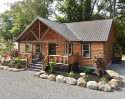 Log cabin home from Salem Structures featuring natural log siding, covered front porch, metal roof, and landscaped stone border on display lot.